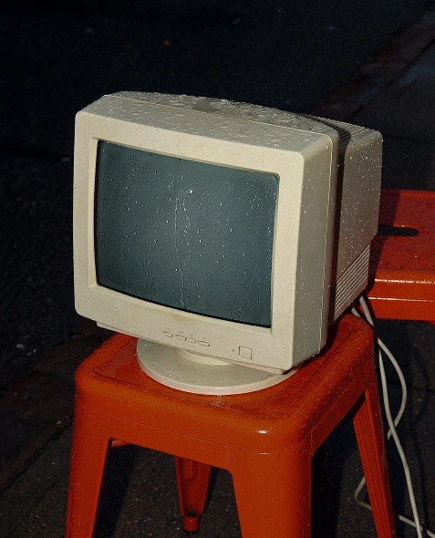 En gammal datorskärm, täckt av vattendroppar, står på en orange pall utomhus