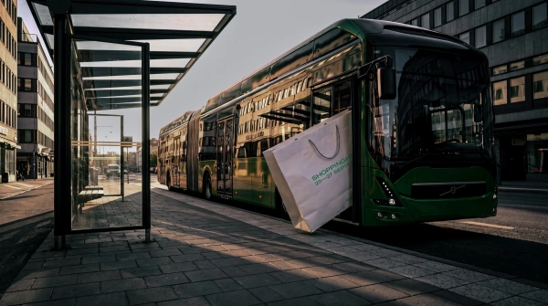 En massiv shoppingpåse med texten "Shoppinghelg 26—27 september" sitter fastklämd i dörrarna på en grön stadsbuss.
